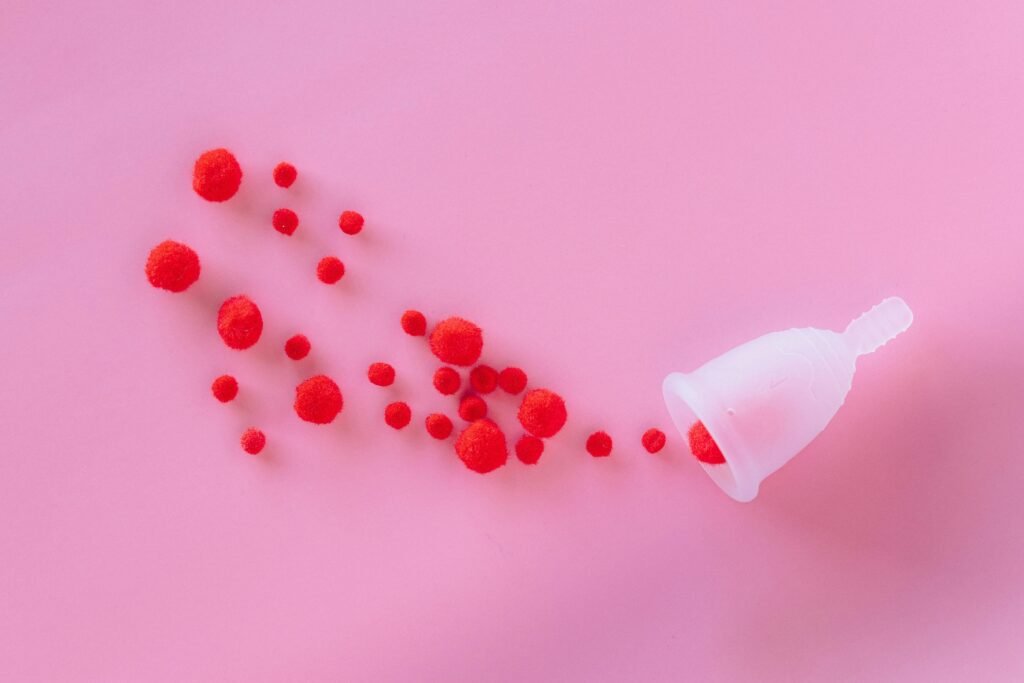 An artistic top-down view of a menstrual cup with red wool balls on a pink background.
