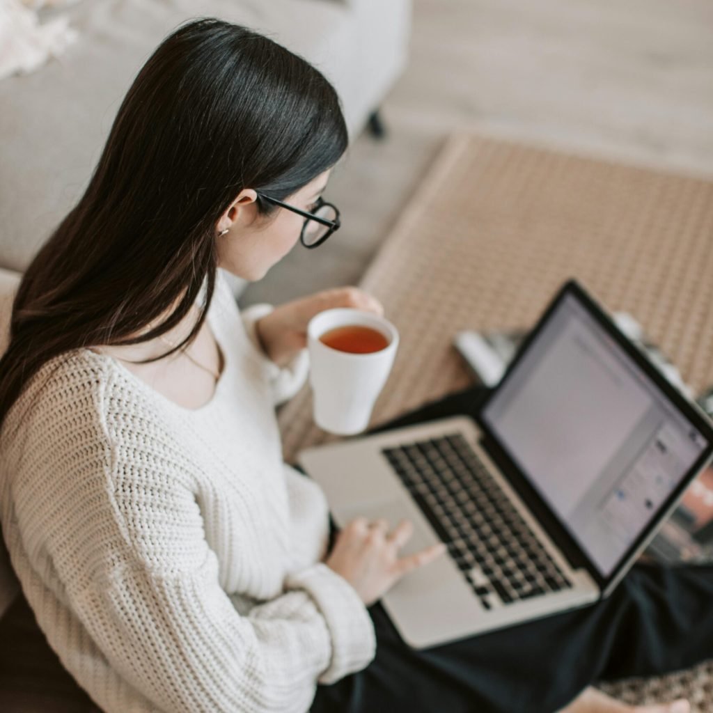 Adult woman in cozy sweater works on laptop while enjoying a hot drink indoors.